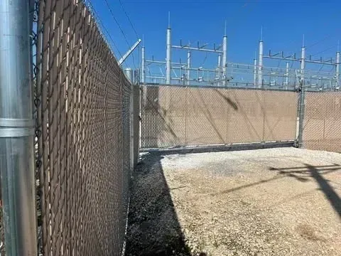 Chain-link fence with brown privacy slats; gate; gravel; power substation in the background under blue sky.