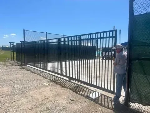 Man standing near an open black metal gate on a gravel surface. A chain-link fence and blue sky are in the background.