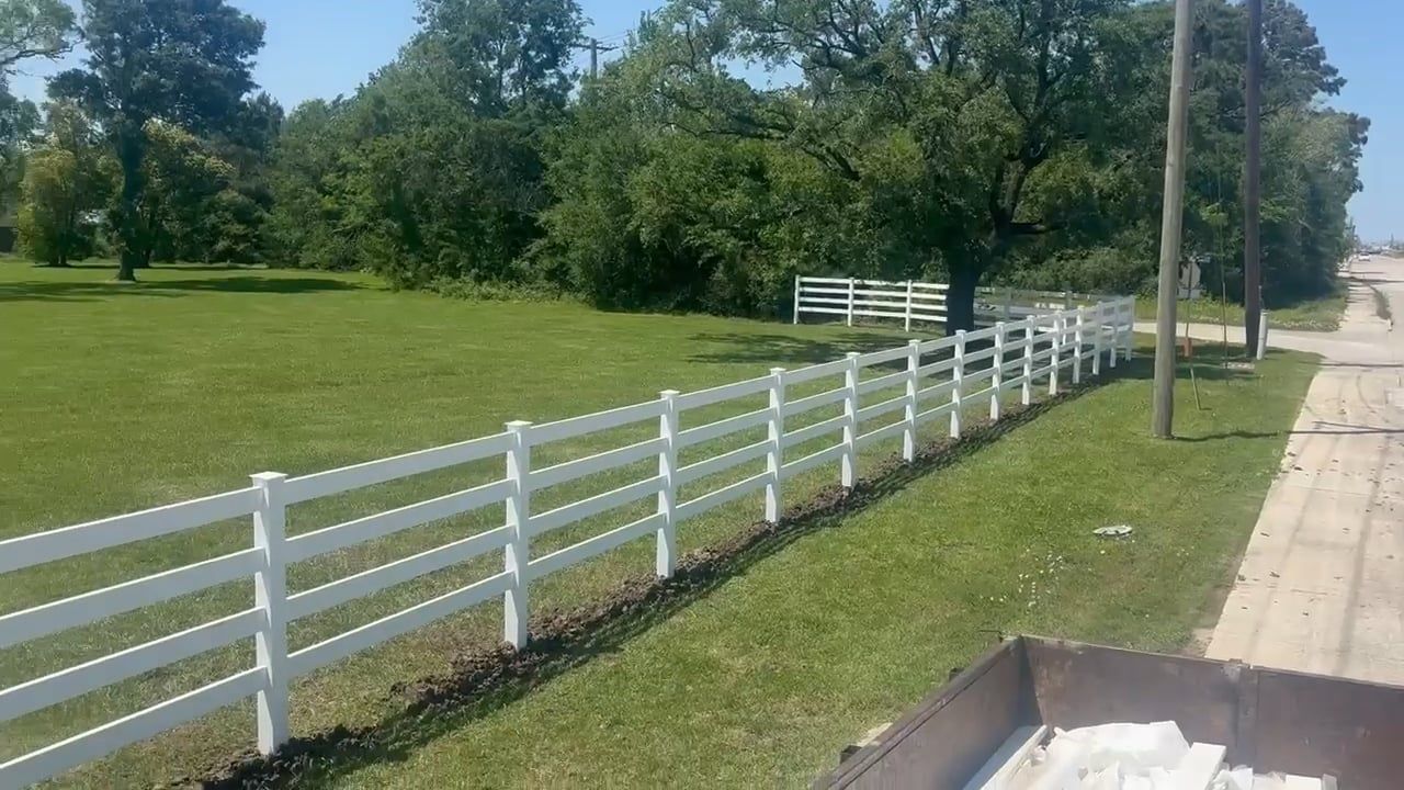 White fence bordering a grassy area with trees and a road in the background. Sunny day.