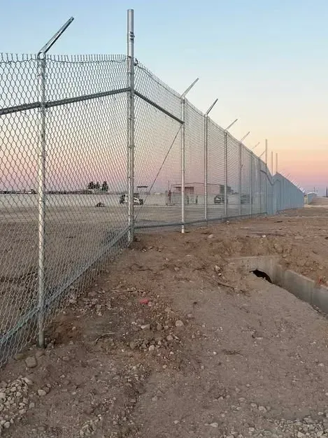 Chain link fence with barbed wire on top, in a dirt field. Evening sky in the background.