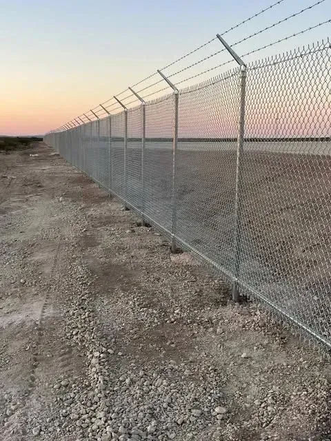 Chain-link fence topped with barbed wire, runs along a gravel path against a dusky sky.