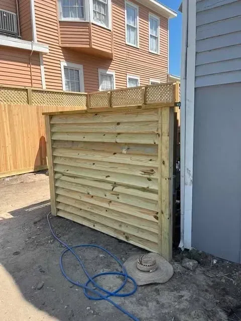 Wooden enclosure against a building; features horizontal slats, lattice top. Blue hose, concrete pad in foreground.