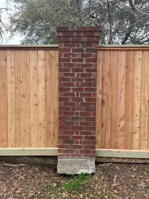 Brick pillar in front of a wooden fence; pillar has a concrete base.