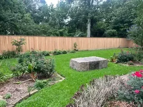 Backyard with wooden fence, green lawn, stone bench, and various plants.