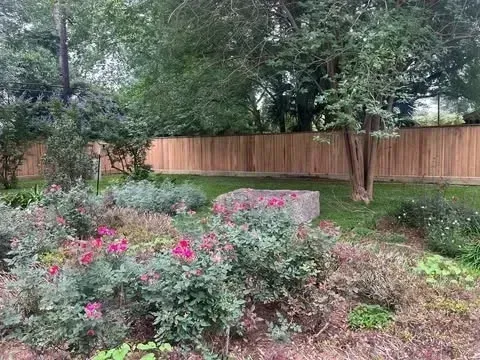 Garden bed with pink flowers and a rectangular stone, with a wooden fence and trees in the background.