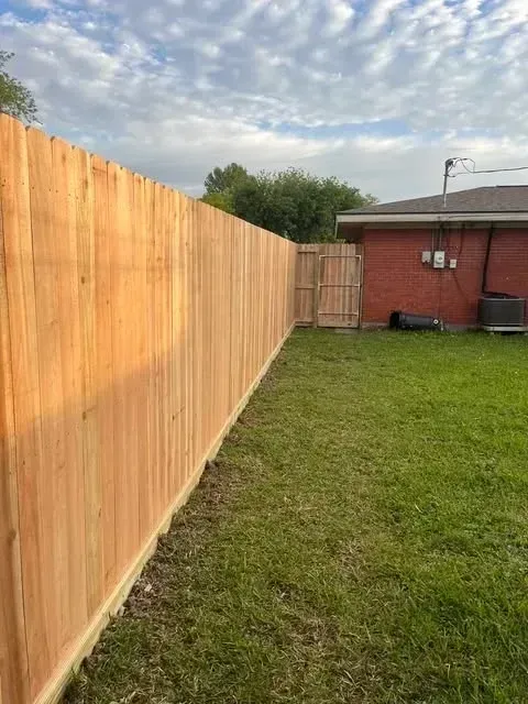 Wooden fence in a grassy backyard with a red brick house and cloudy sky in the background.