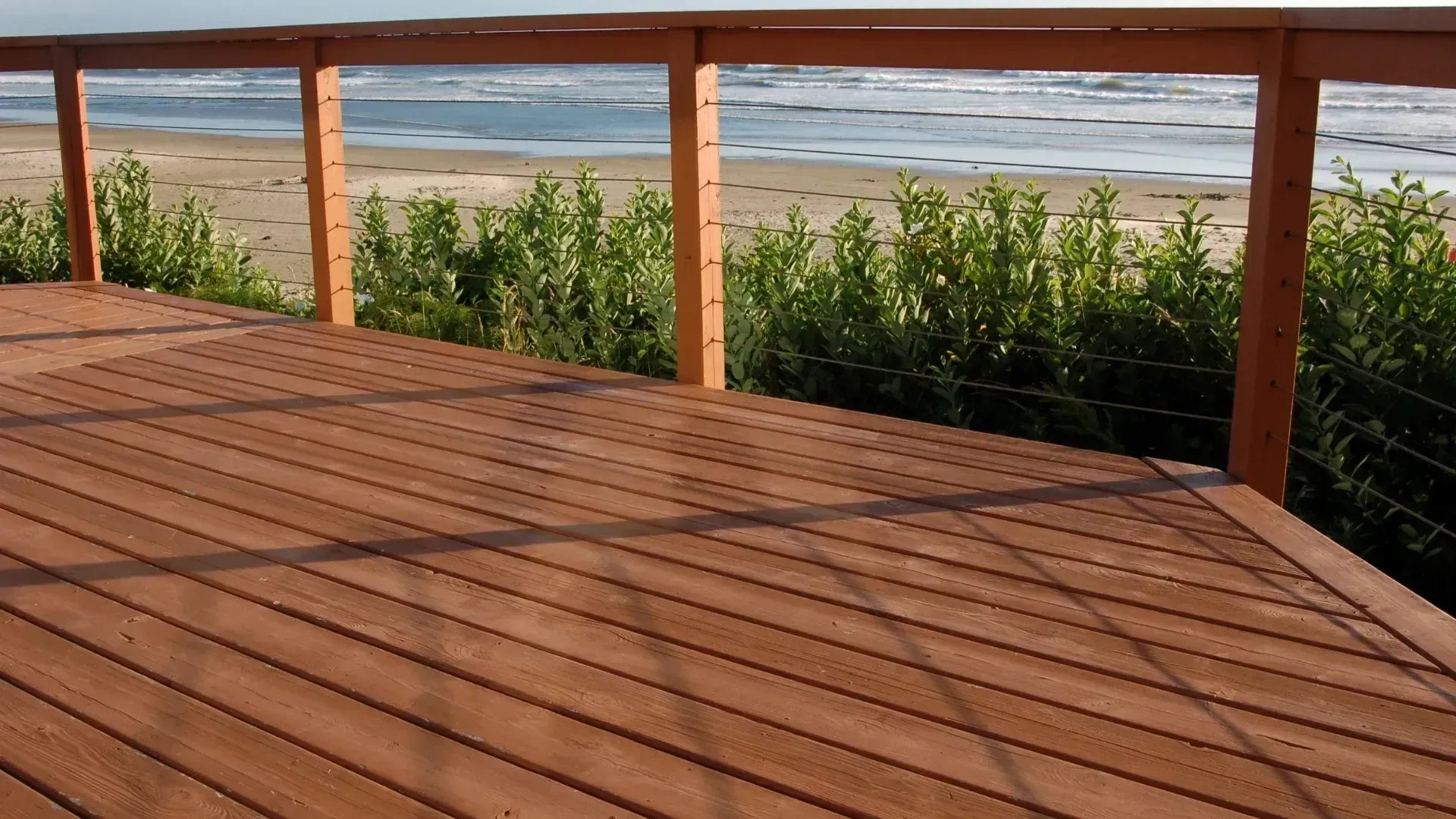 Wooden deck overlooking a beach and ocean with green plants and a wooden railing.