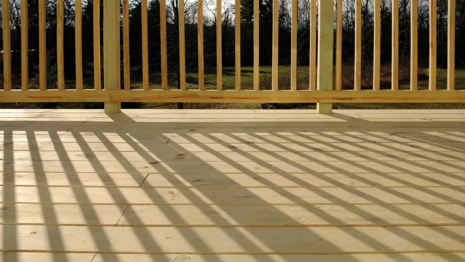 Wooden deck with railing casting shadows on the deck's surface. Trees are visible in the background.