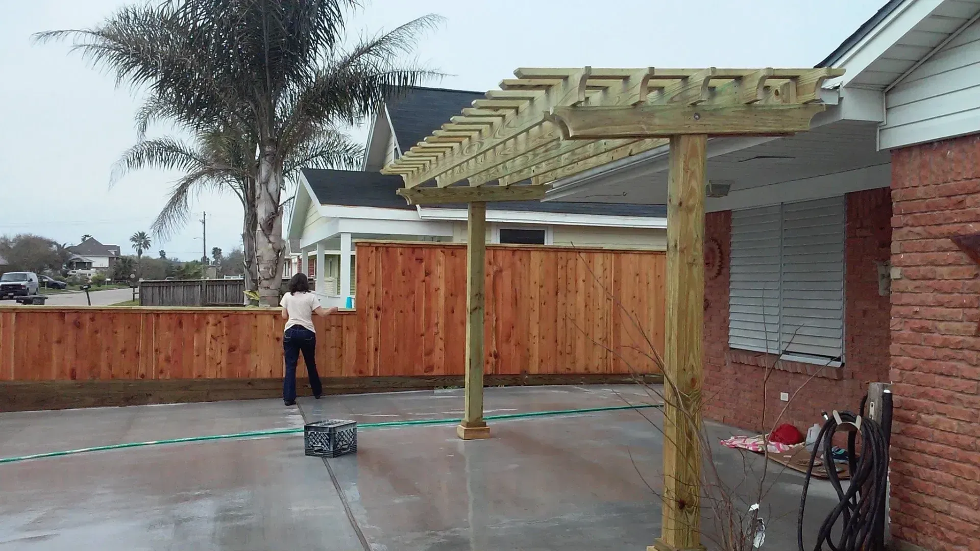 Woman using a power washer on a wet patio. Wooden pergola attached to a brick house and fence.