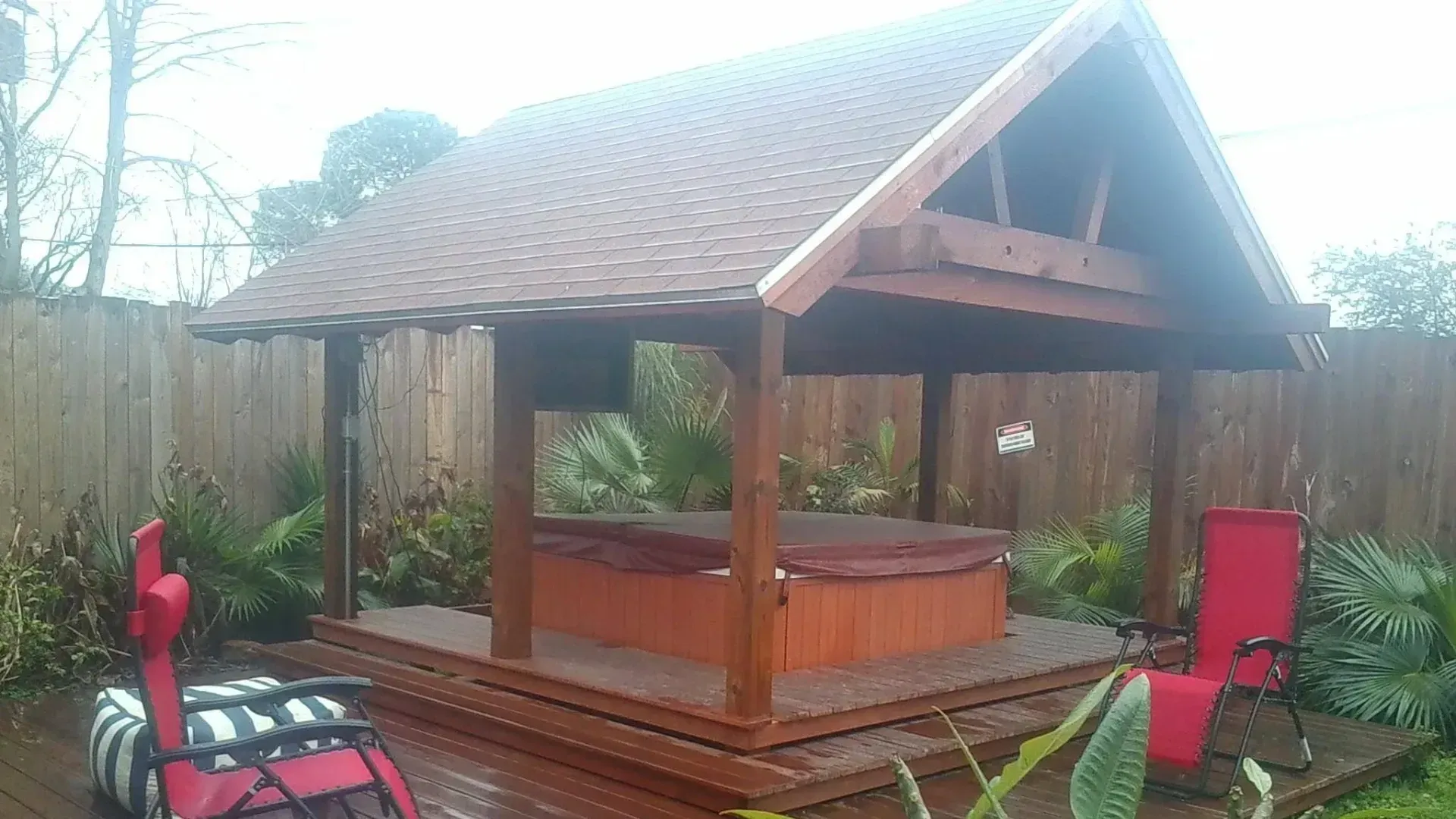 Wooden gazebo with hot tub, red chairs, and a garden setting.
