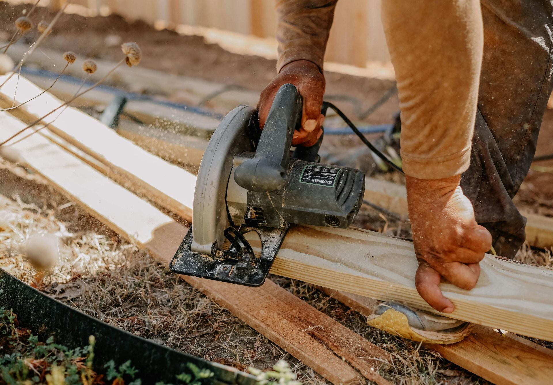 Person using a circular saw to cut wooden planks outdoors, with sawdust flying.