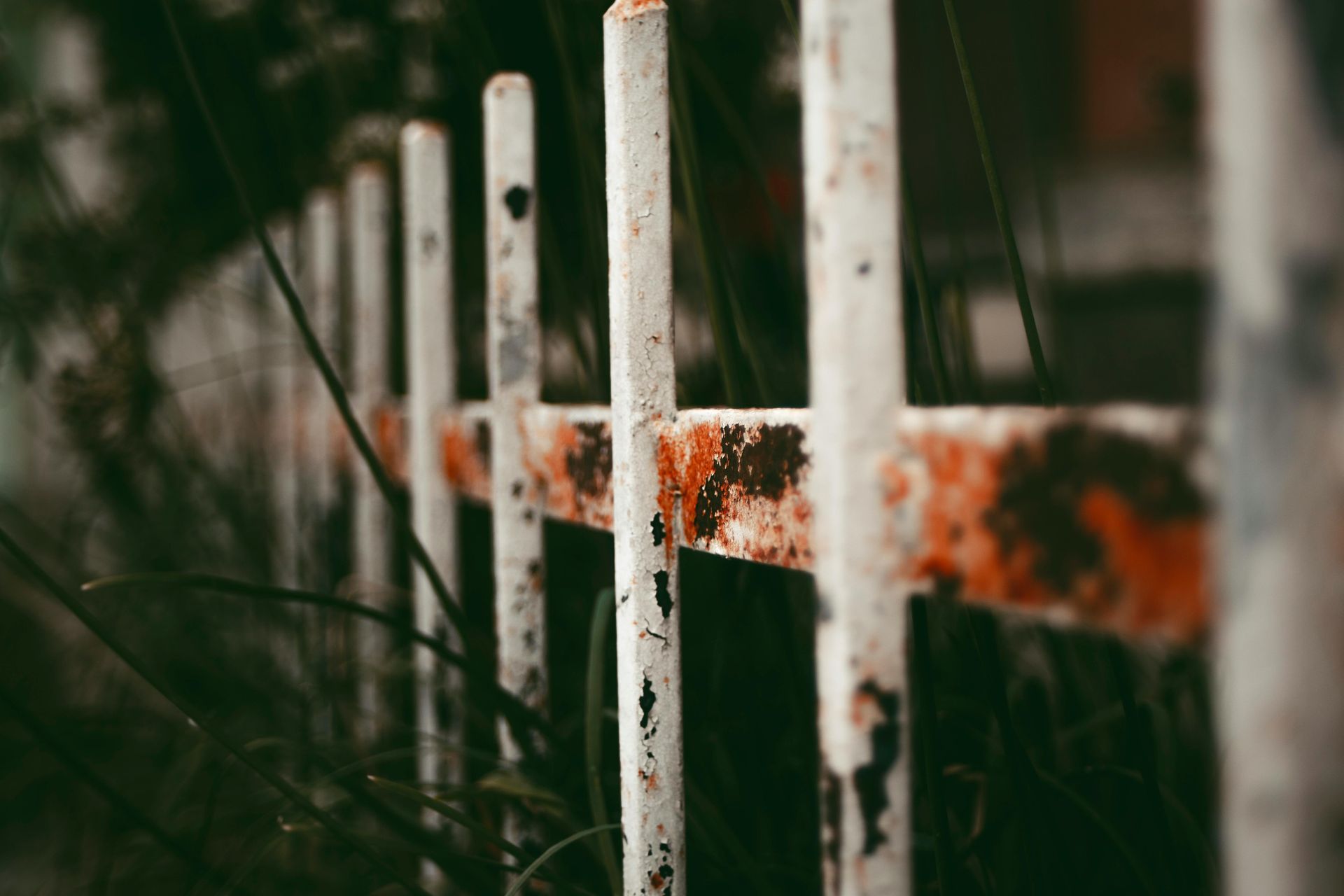 A close-up, angled view of a weathered white metal fence with orange rust patches, set against a blurry, dark background.