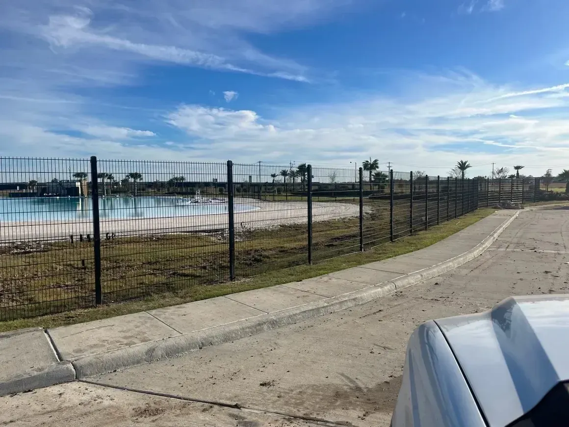 Black fence bordering a light-colored pool and concrete path on a sunny day.