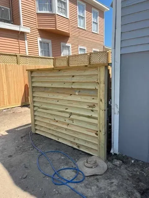 Wooden enclosure with horizontal slats and latticework top, next to a building. Blue hose and hat on the ground.