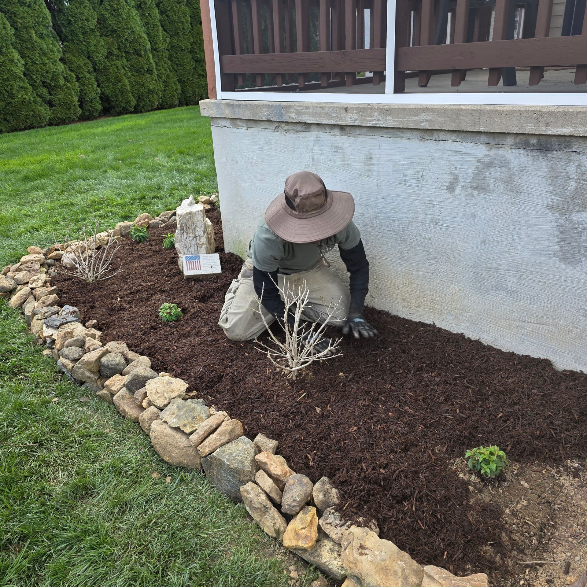 Person planting in a flower bed near a concrete foundation. They wear a hat, gloves, and khaki pants.