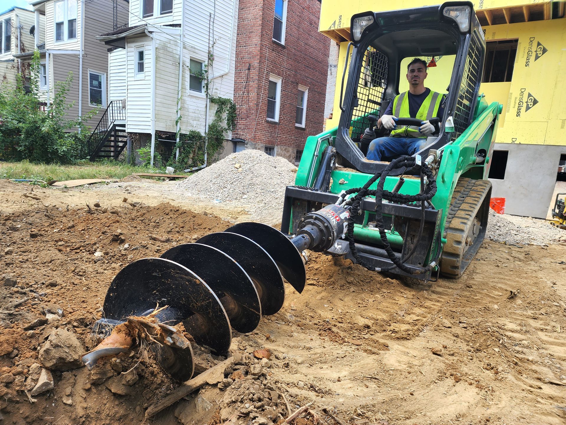 Man operating a green skid steer with an auger drilling into the ground at a construction site.