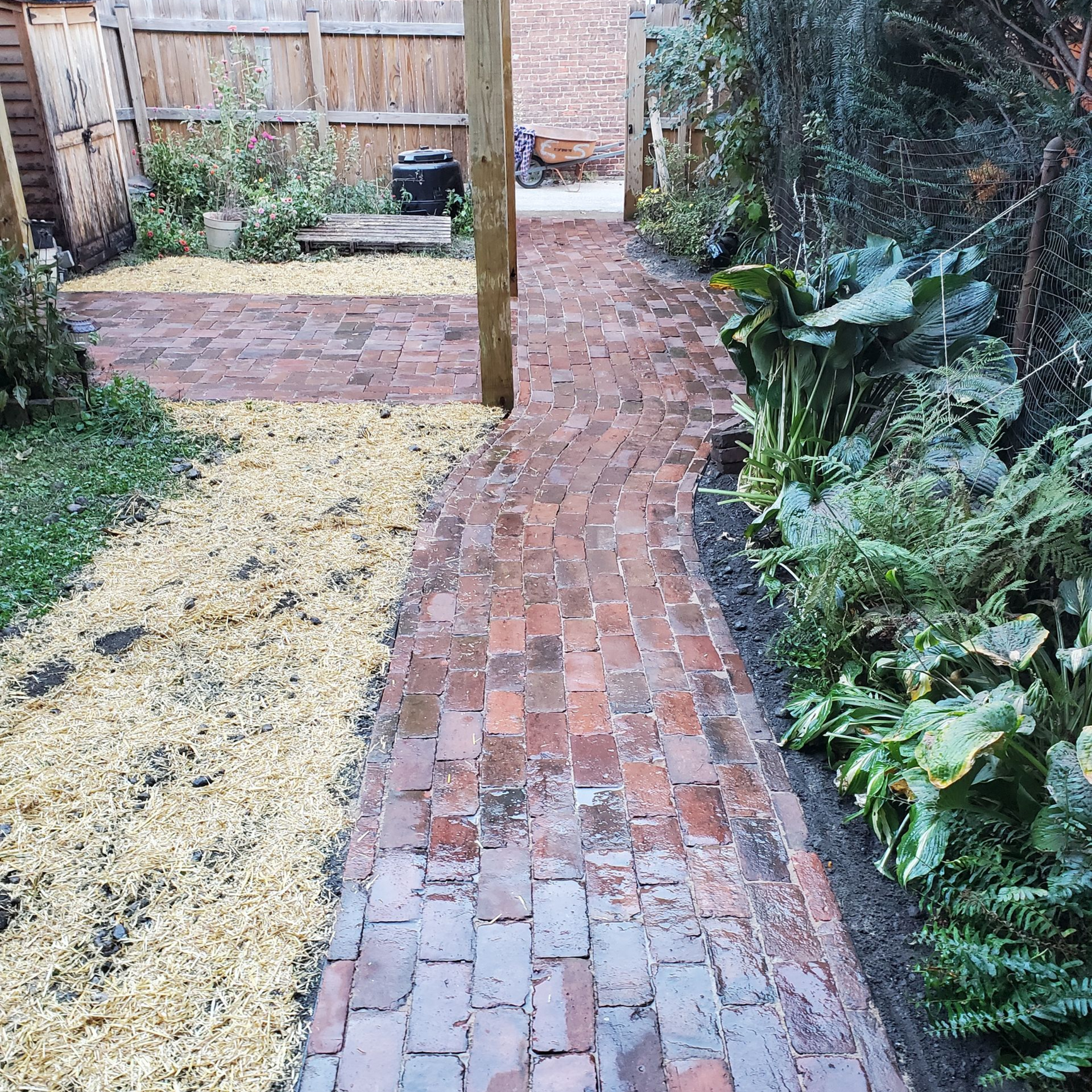 Brick path in a garden, bordered by plants and wood post. A side path has yellow mulch.