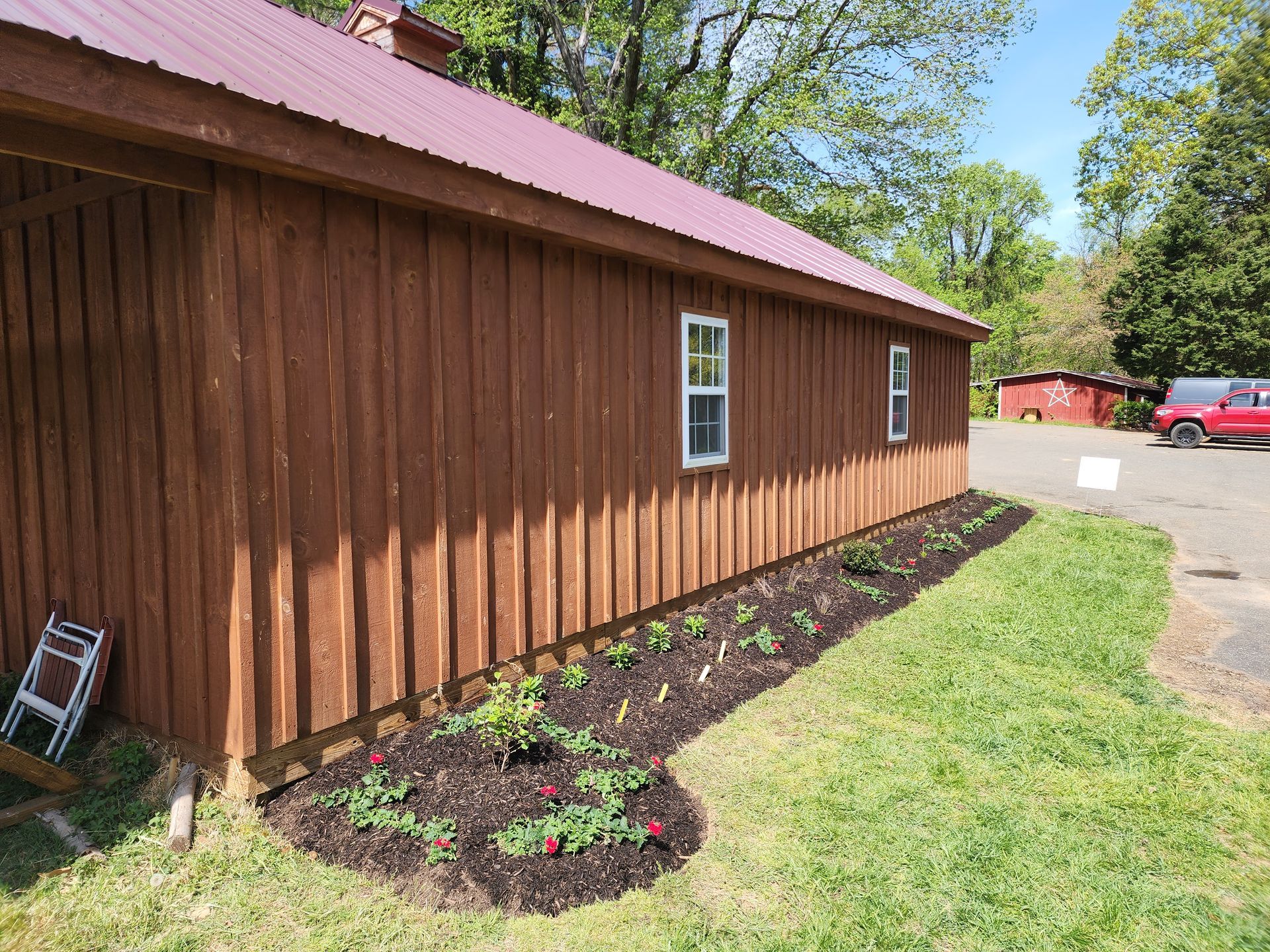 Brown barn with a red roof, two white windows, and a flower bed along the base.