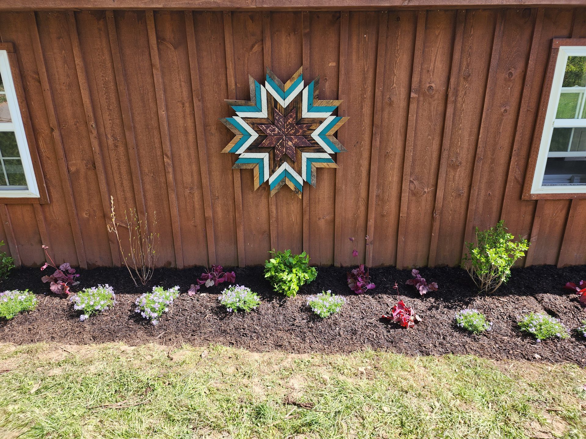 Brown wooden wall with a teal and brown starburst decoration and a flower bed with various plants in front.