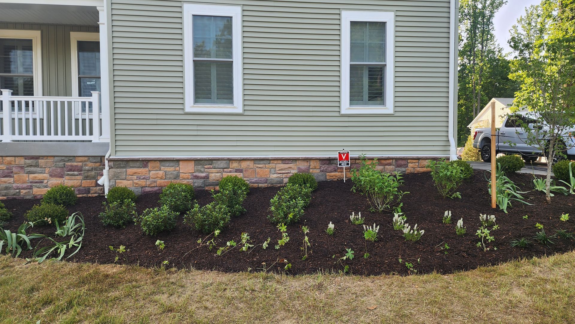 House with flower bed, green siding, stone facade. Boxwood shrubs, dark mulch, sunny.