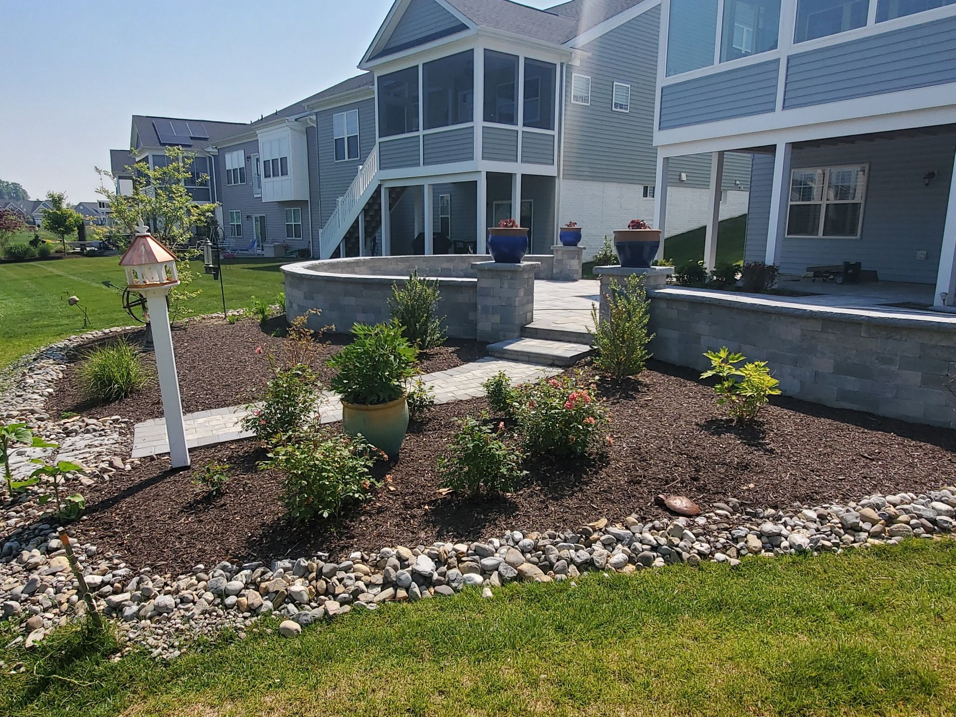 Backyard landscaping with stone patio, flower beds, and house in the background.