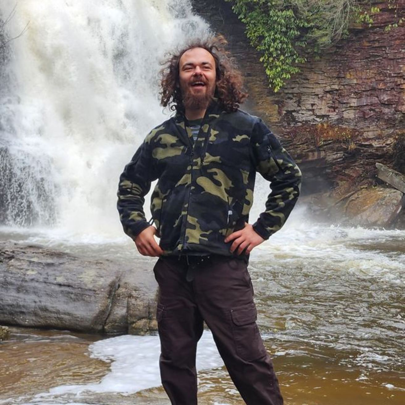 Man with curly hair smiles in front of a waterfall; he wears a camo jacket and brown pants.