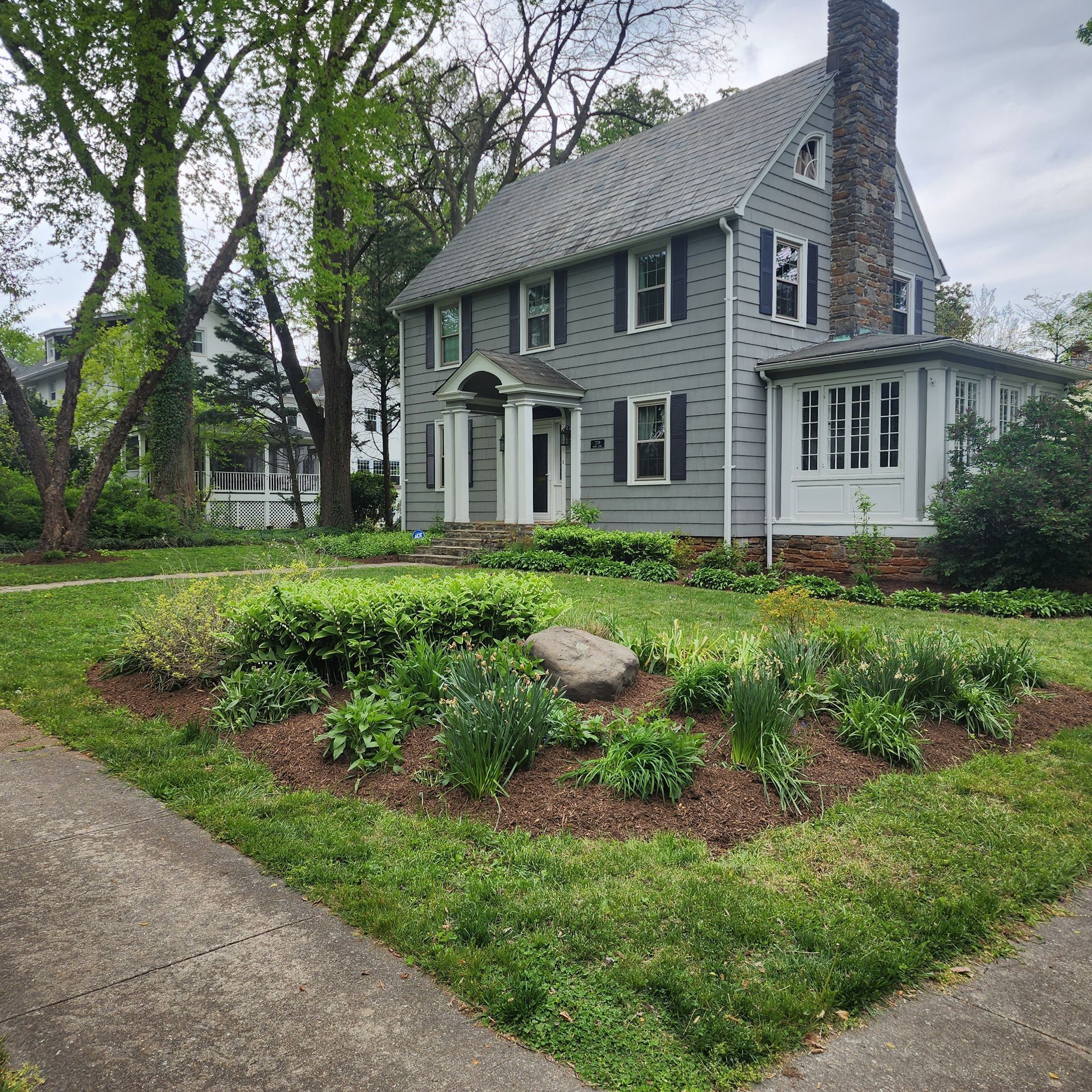 Gray house with a small garden in front. A large stone sits in the center of the flower bed.