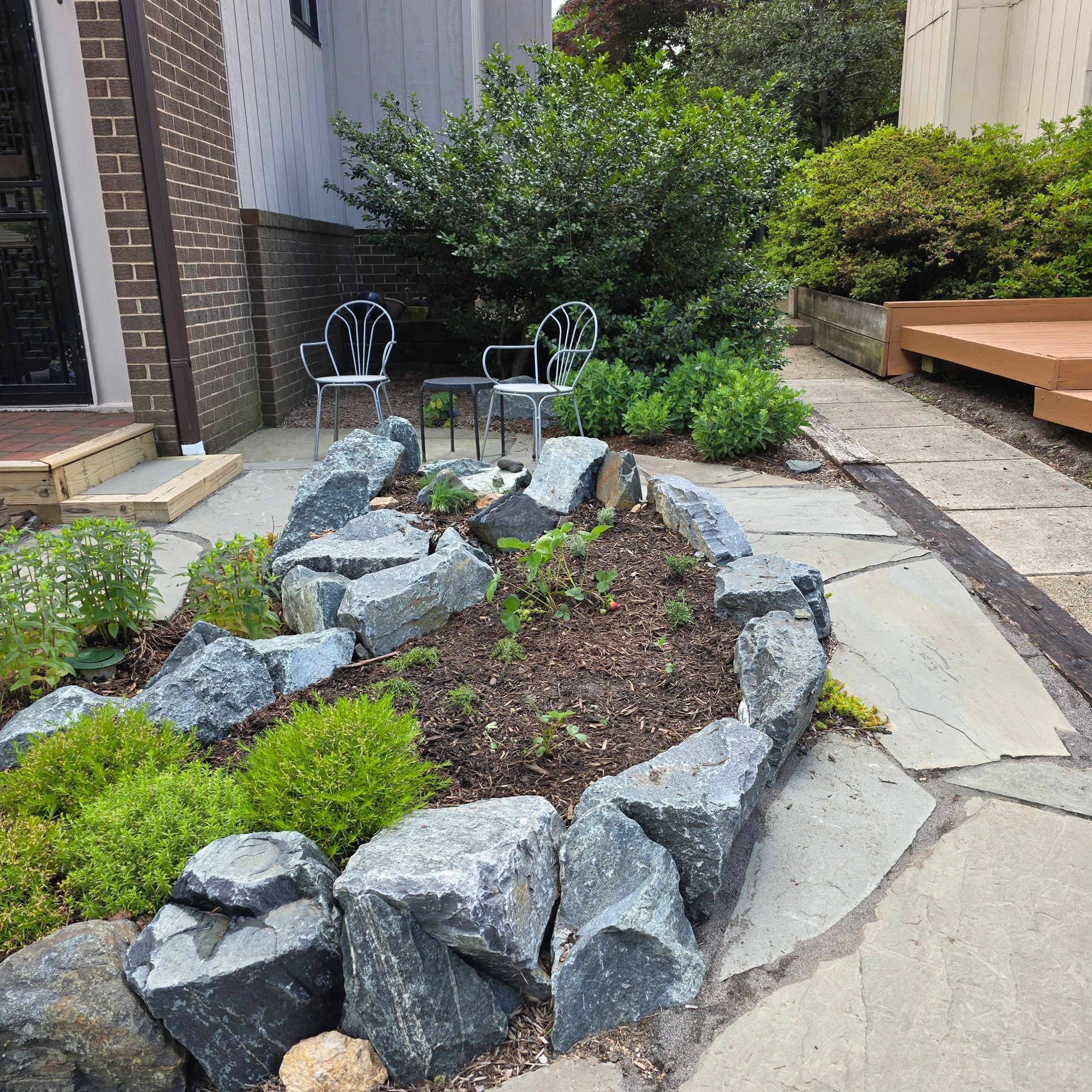 Stone-lined garden bed with rocks and plants. Two chairs and table in the background.