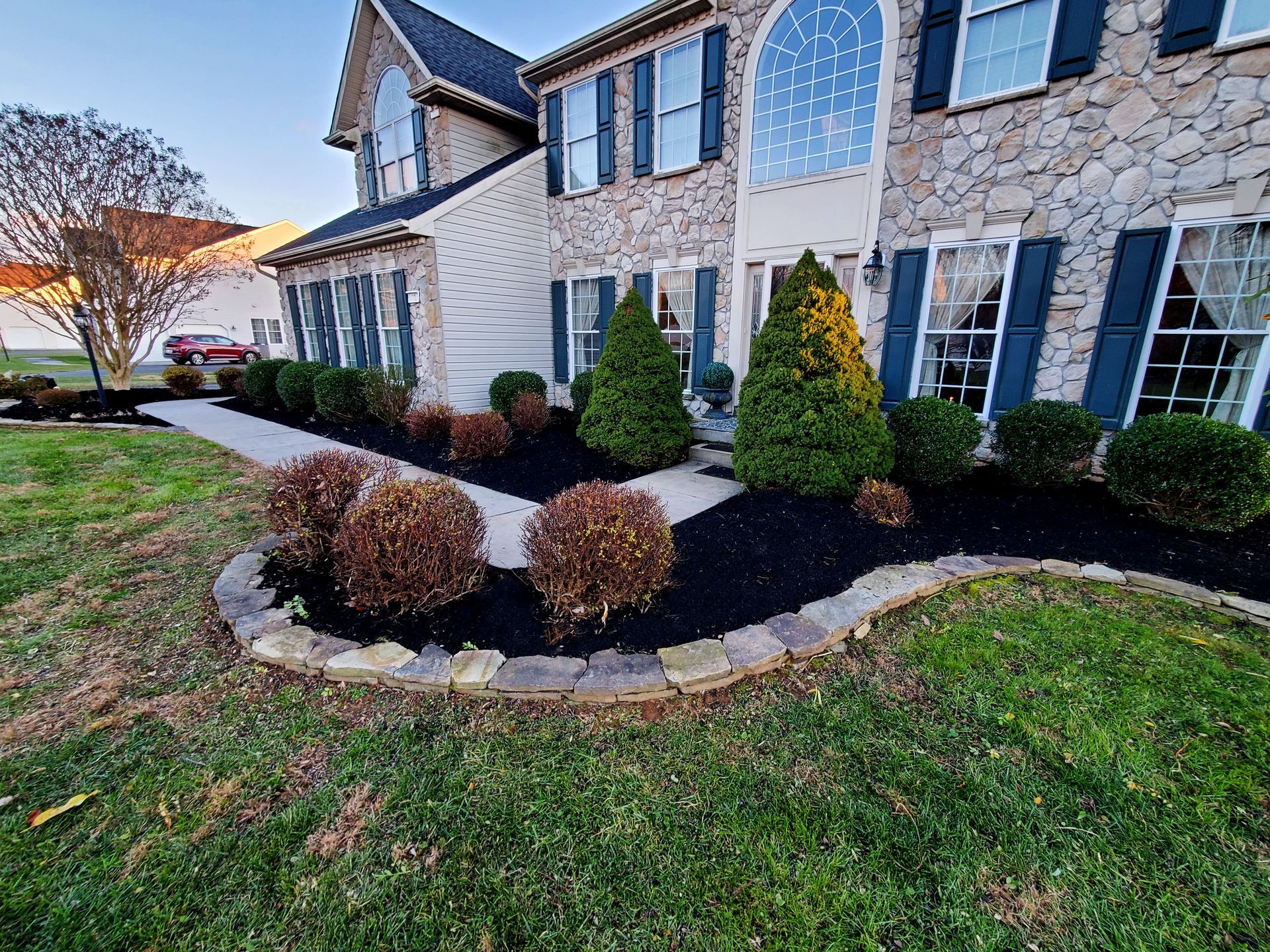 Two-story house with stone facade, neat landscaping, dark mulch, manicured bushes, and a sidewalk.