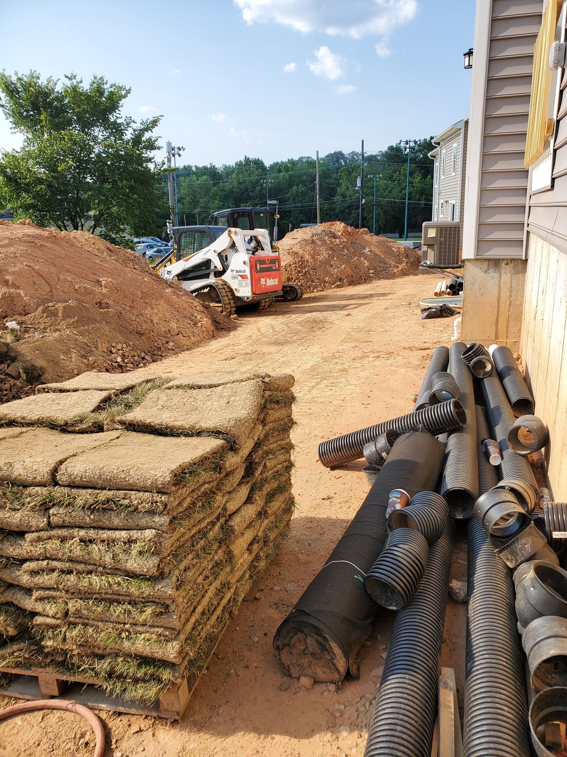 Construction site: sod on pallets, Bobcat digging earth, drainage pipes against building.