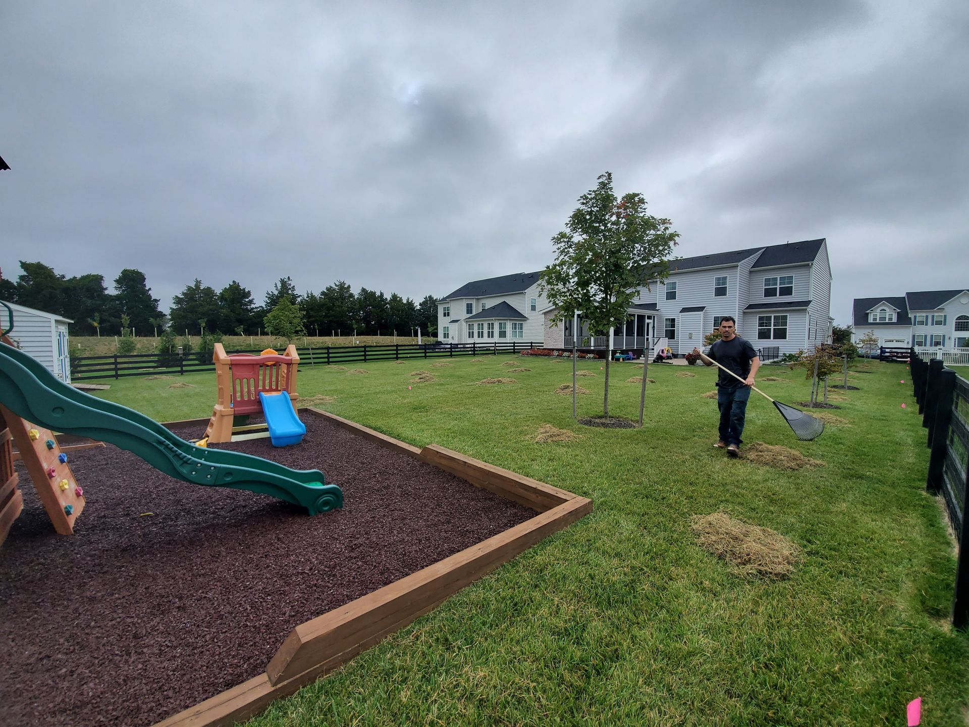 Man raking grass in backyard near a playground and trees, with houses in the background under a cloudy sky.