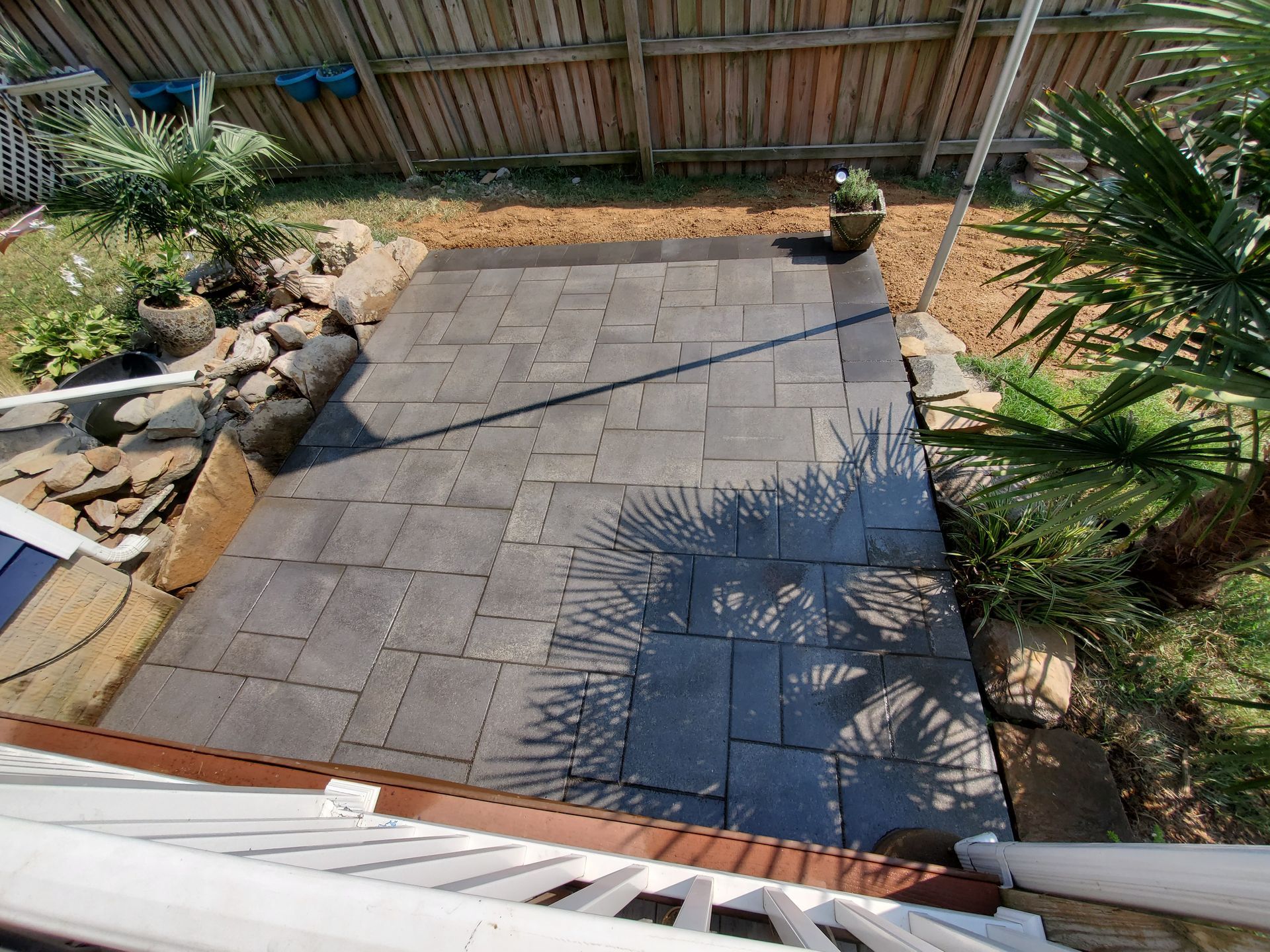 Gray stone patio in a backyard, surrounded by greenery and wooden fence, viewed from above.