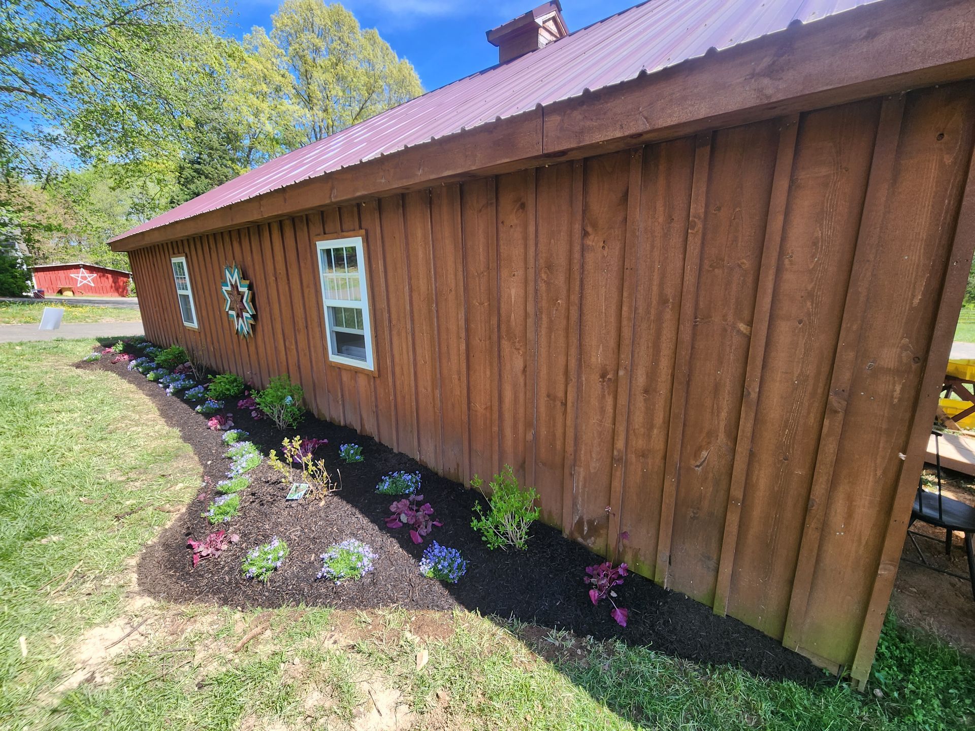 Brown barn with a red roof, bordered by a flower bed with mulch, plants, and green grass.
