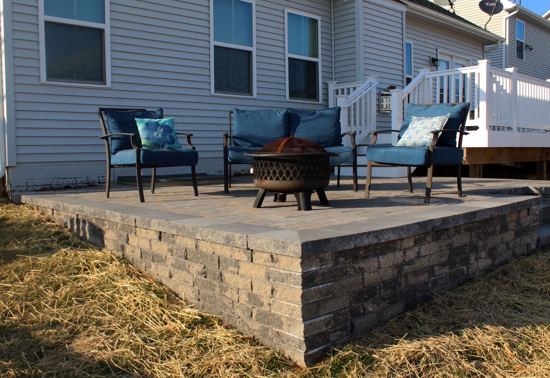 Outdoor patio with blue furniture and fire pit. Brown and grey brick retaining wall with brown grass.