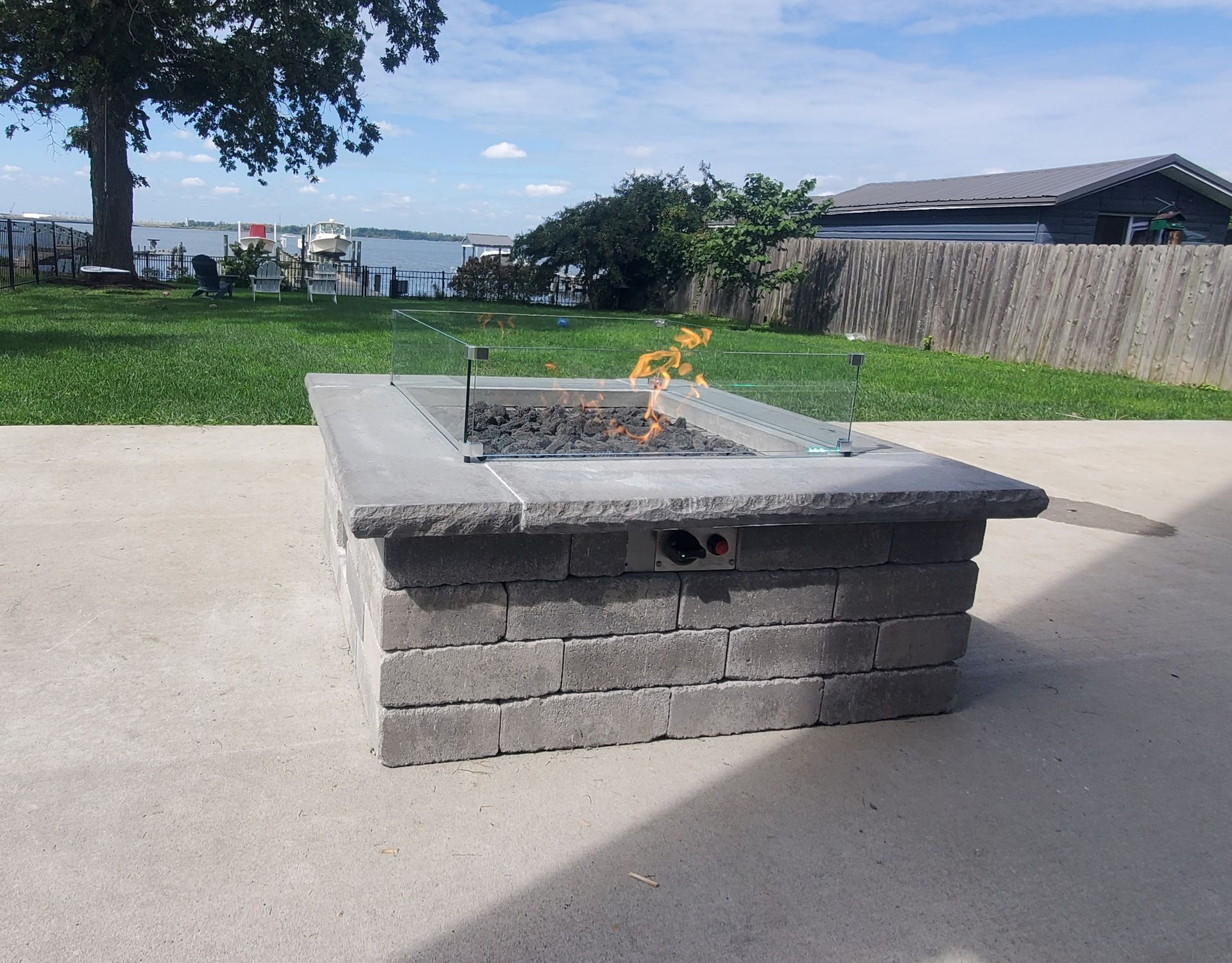 Brick fire pit with a small flame on a concrete patio, overlooking a lake.