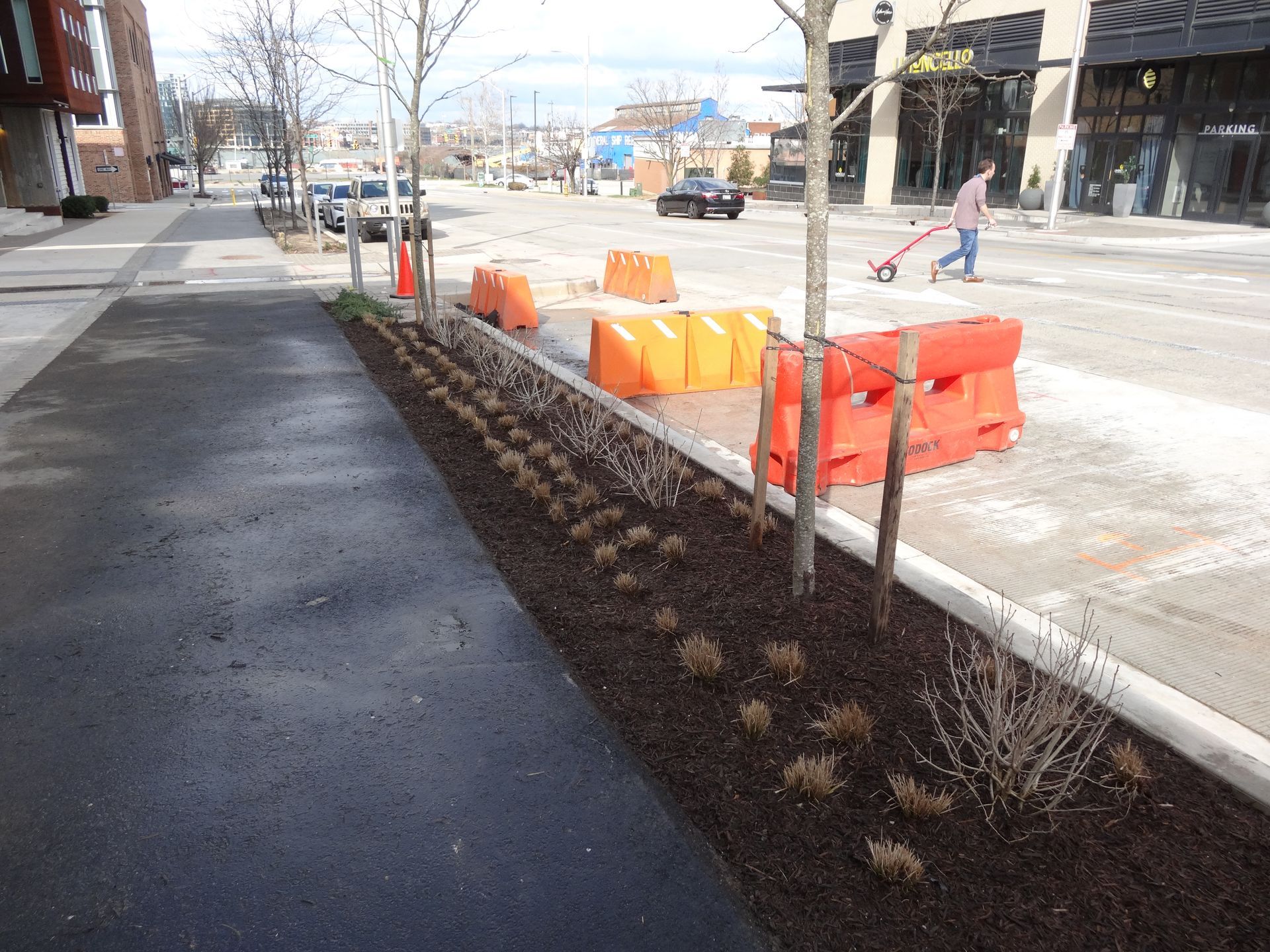 Asphalt sidewalk with a planted bed of brown mulch and dormant plants, orange safety barriers, and a person sweeping.