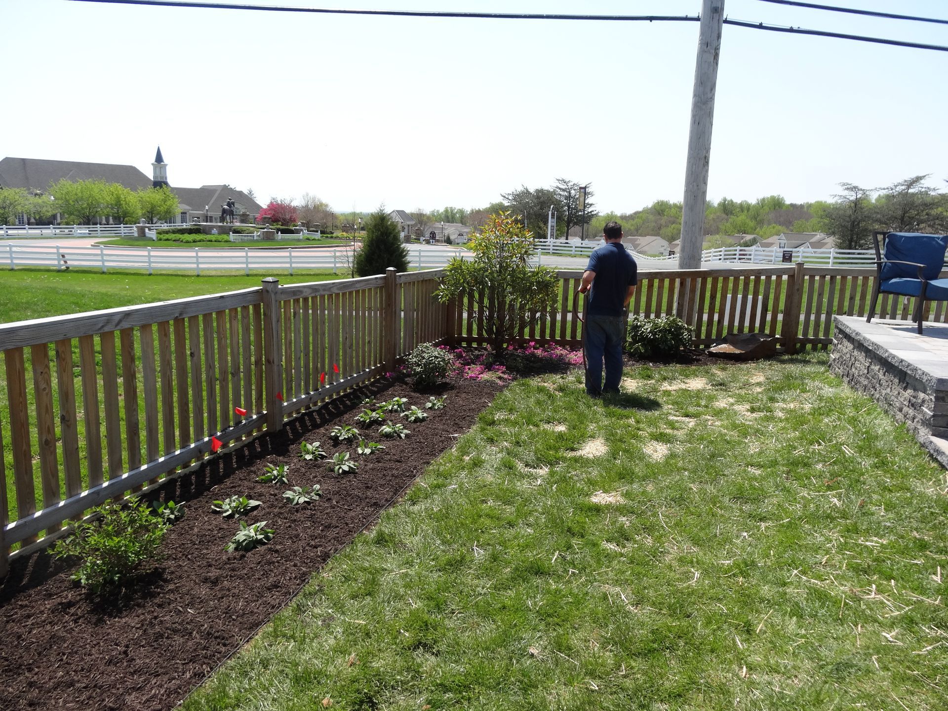 Man standing by a wooden fence, looking at a flower bed and grassy yard on a sunny day.