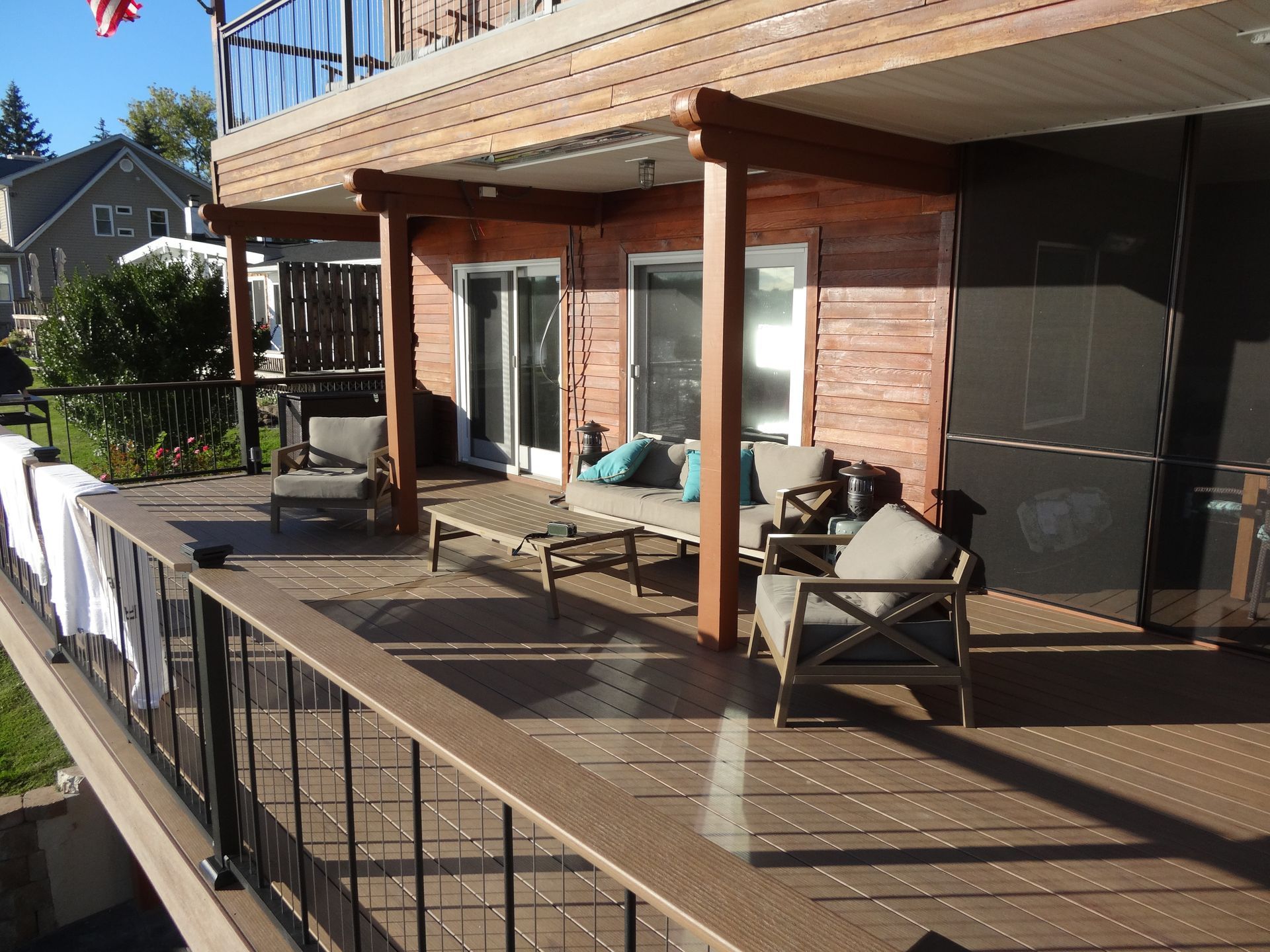 Deck with furniture, shaded area, overlooking a yard and a house. Brown deck, wood posts, and seating.