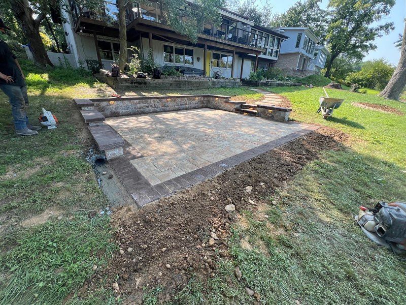 A paved patio with retaining walls and a house in the background. Construction materials and a worker are visible.