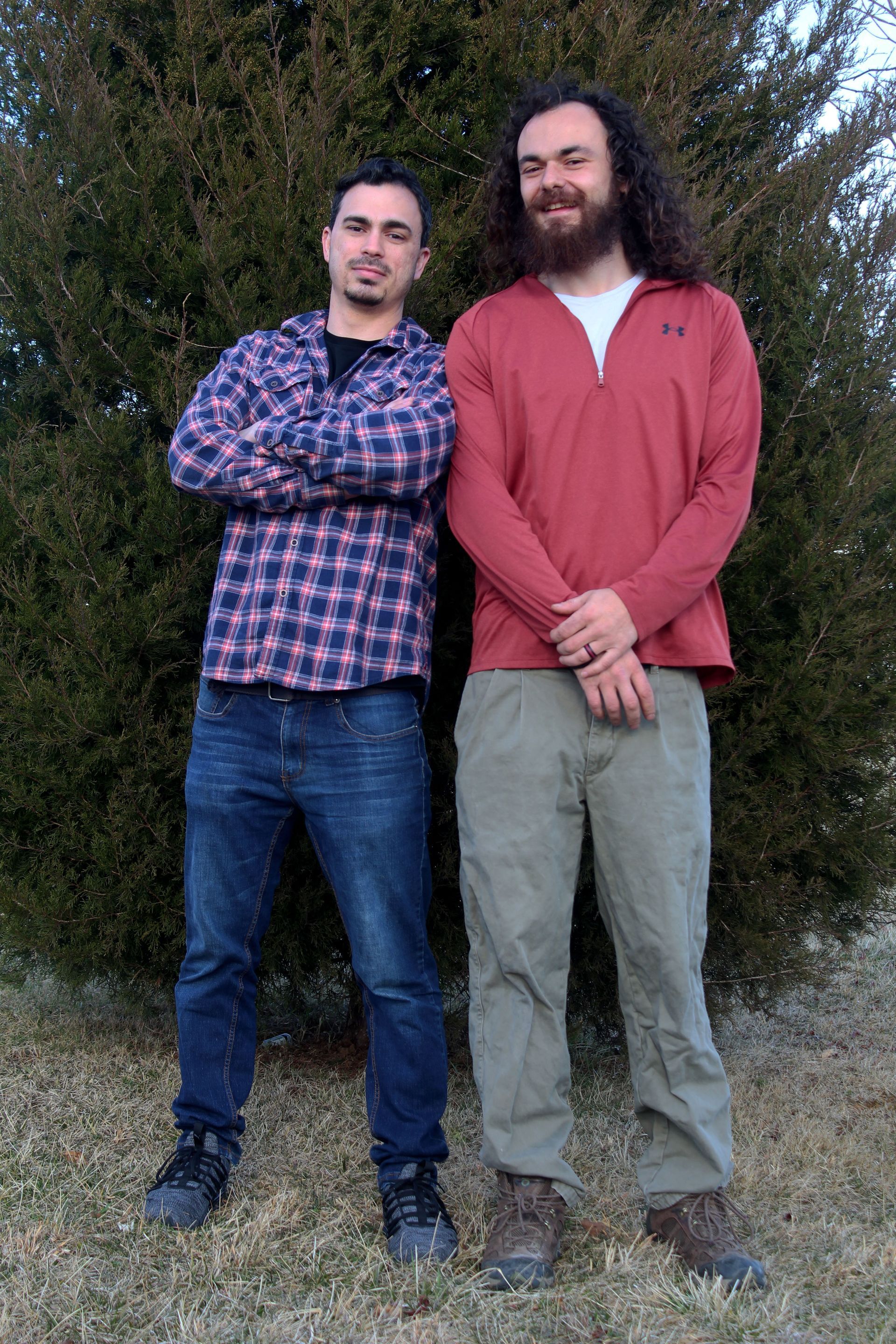Two men pose outdoors: one in a plaid shirt, jeans, and crossed arms; the other in a red shirt and green pants.