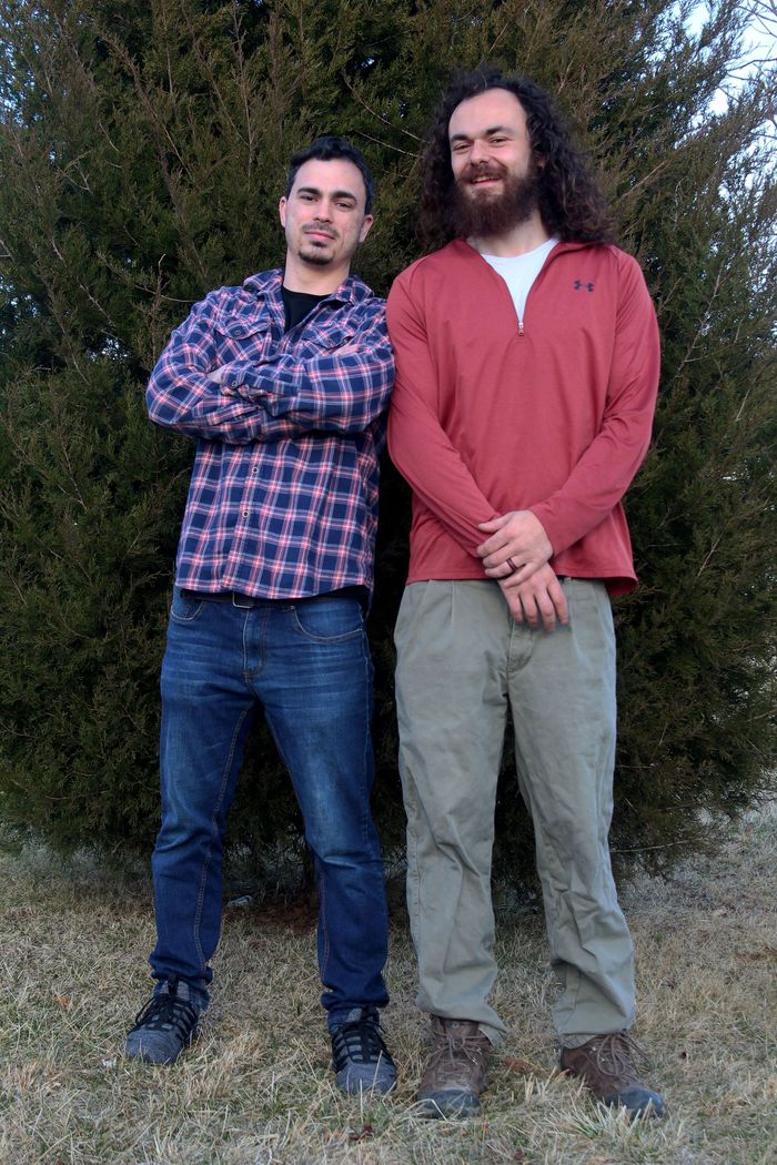 Two men pose outdoors: one in a plaid shirt, jeans, and crossed arms; the other in a red shirt and green pants.