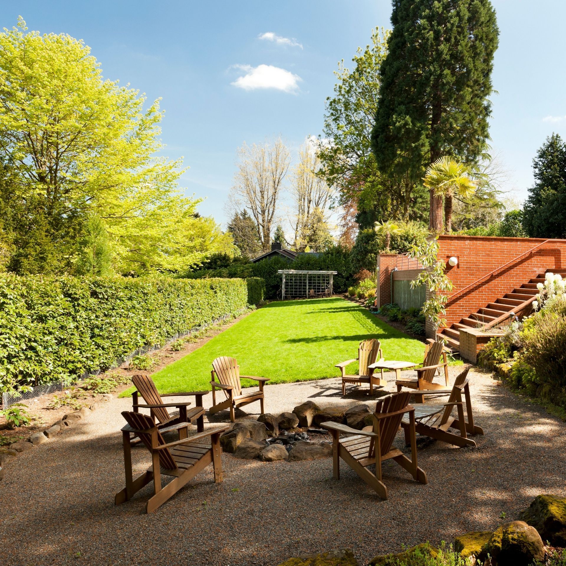 A garden with a fire pit, wooden chairs, and a grassy lawn, surrounded by trees and hedges.