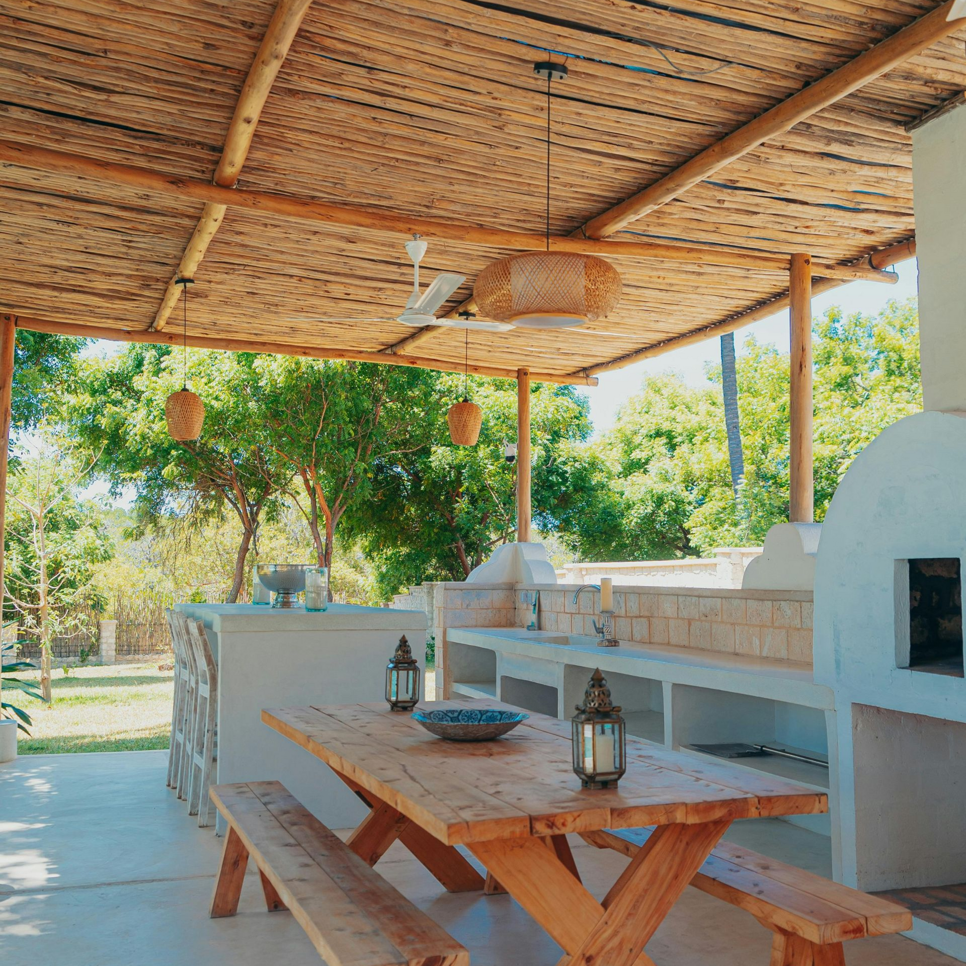 Outdoor dining area with wooden table, benches, and a pizza oven under a thatched roof.
