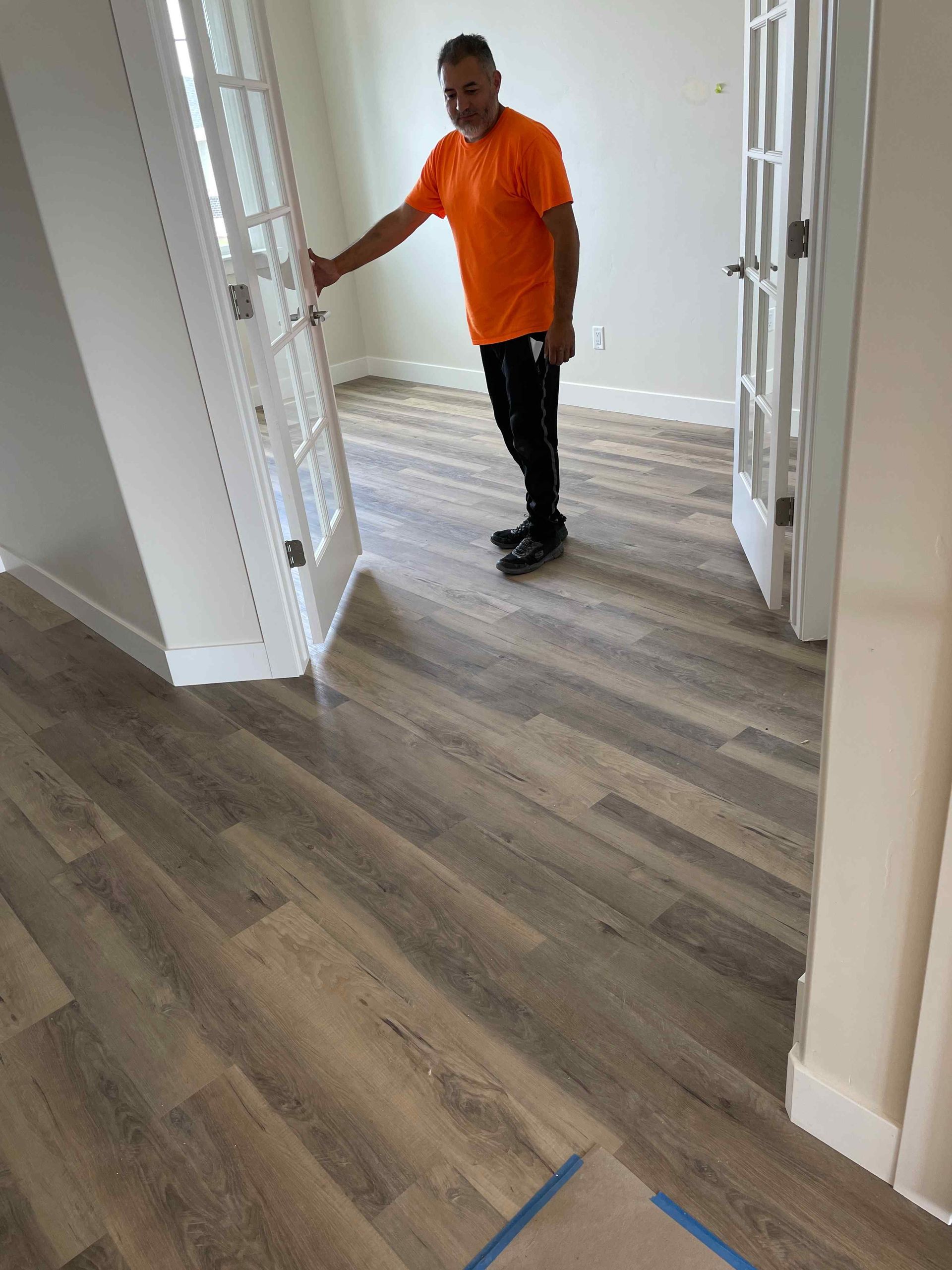 A man in an orange shirt is standing on a wooden floor in a room.