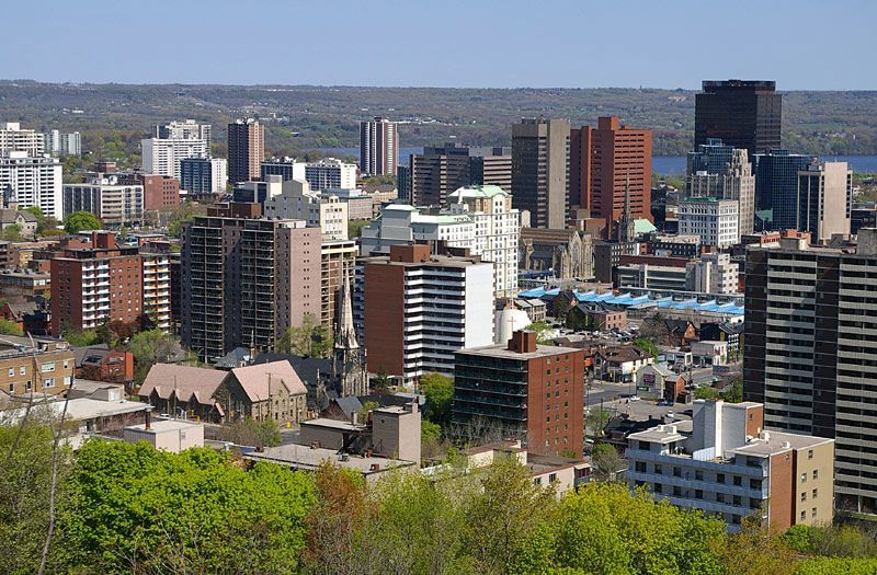 A cityscape of Hamilton, Ontario, featuring various apartment buildings and offices with a treeline in the foreground.