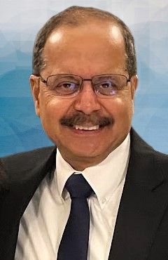Man wearing glasses, dark suit, and tie, smiling.