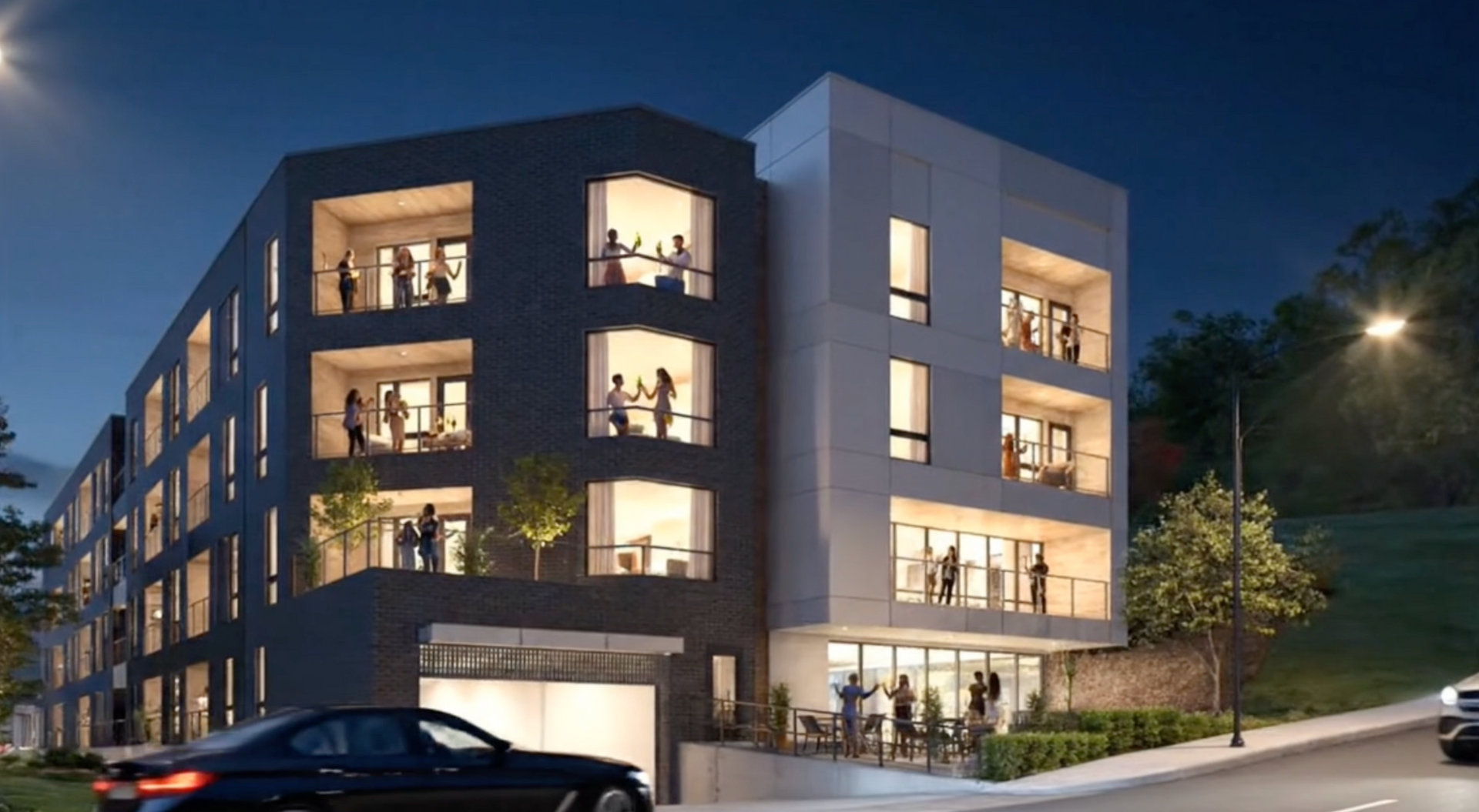 Modern apartment building at night; people on balconies, lights on, car parked in front.