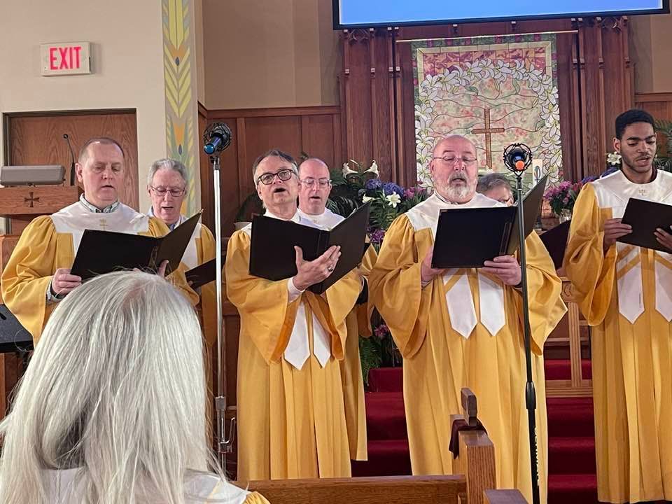 A group of men singing in a church with an exit sign in the background