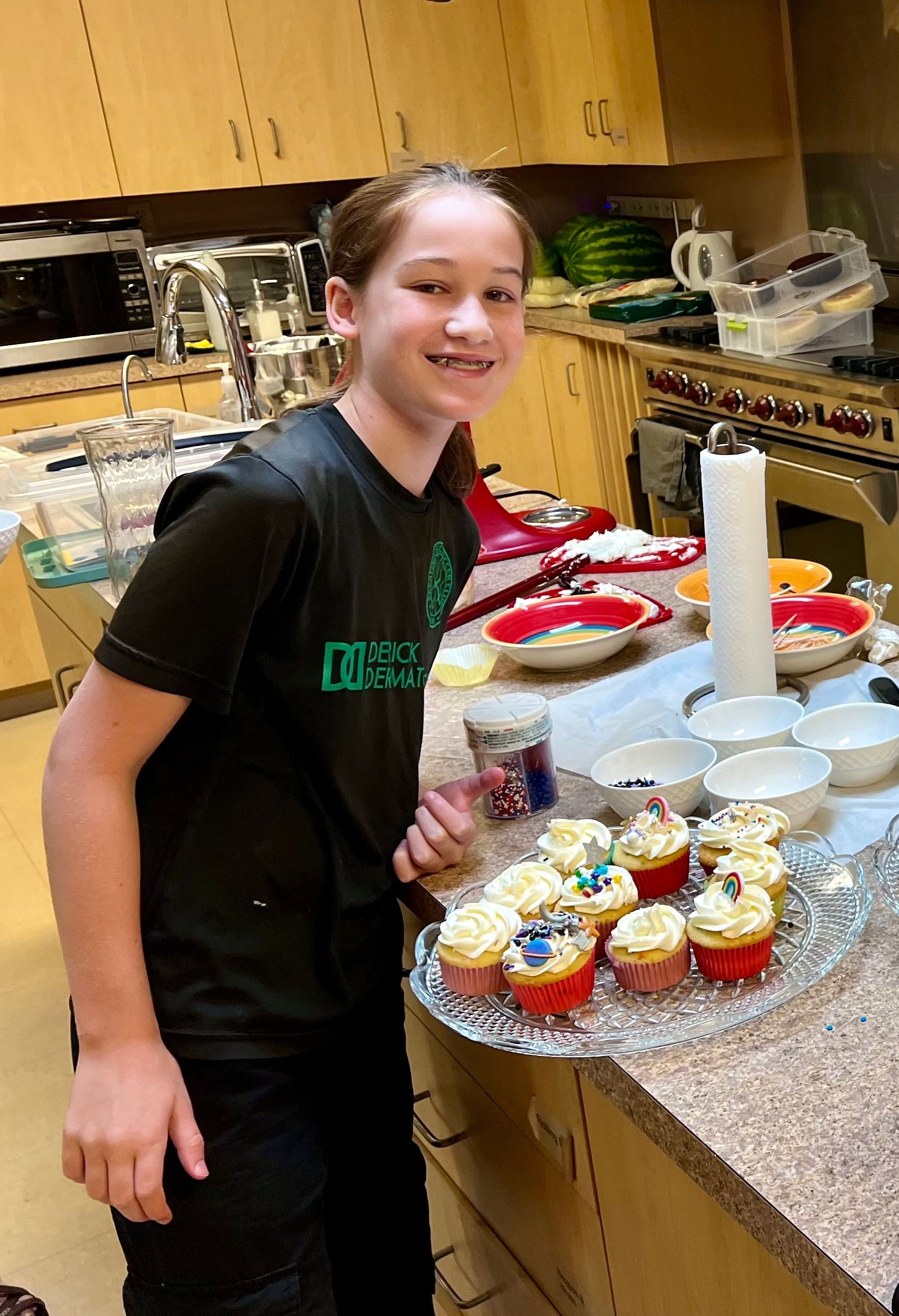 A young girl is standing in a kitchen holding a plate of cupcakes.