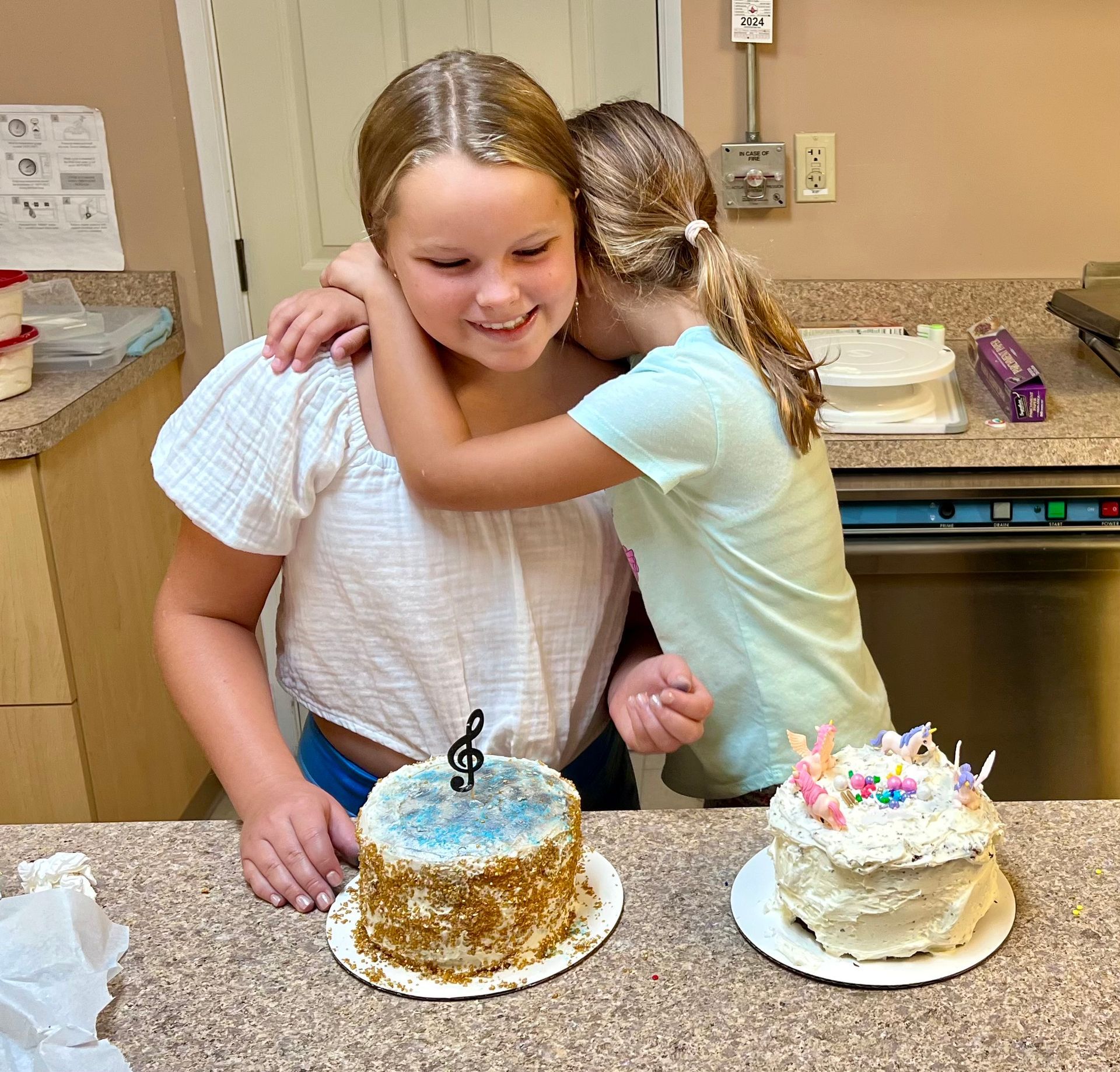 Two young girls are hugging each other in front of two cakes on a counter.